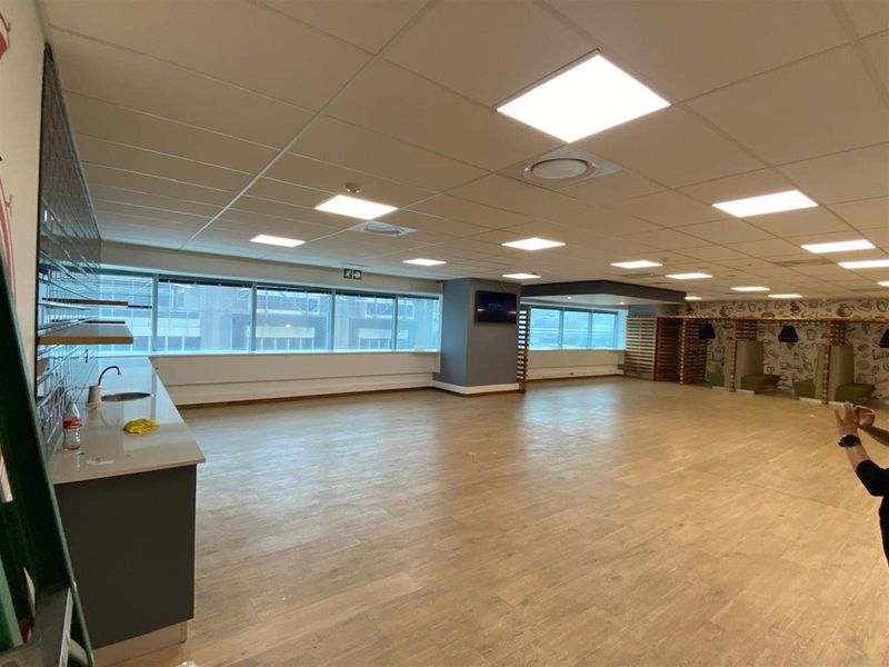 Spacious open-plan office area featuring a kitchenette, large windows for natural light, suspended ceiling tiles, and wood-look flooring.