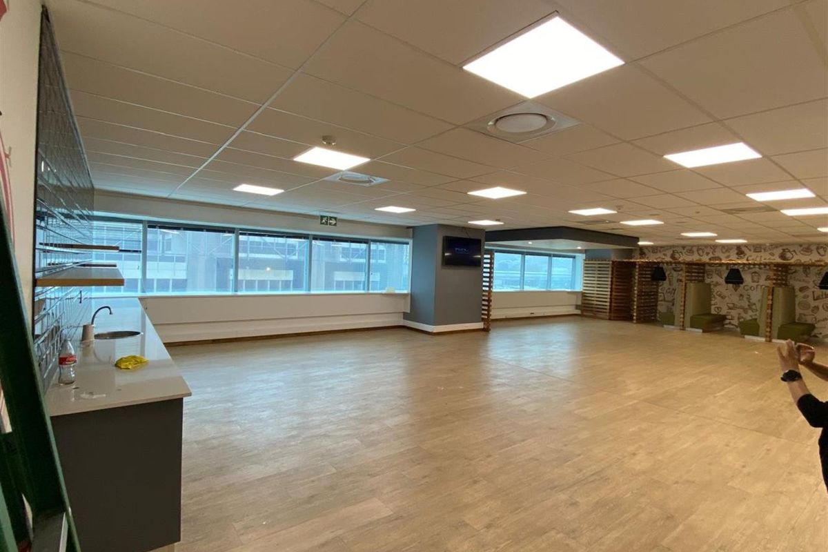 Spacious open-plan office area featuring a kitchenette, large windows for natural light, suspended ceiling tiles, and wood-look flooring.