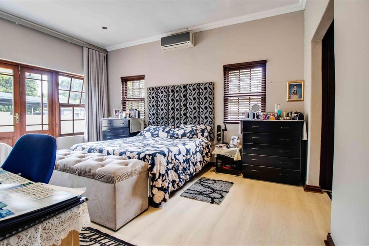 A well-lit bedroom featuring light-toned laminate flooring, wooden window blinds, and a wall-mounted air conditioning unit.