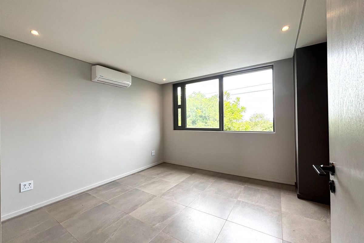 A bright, modern bedroom featuring large windows, neutral tiled flooring, recessed ceiling lighting, and a wall-mounted air conditioning unit.