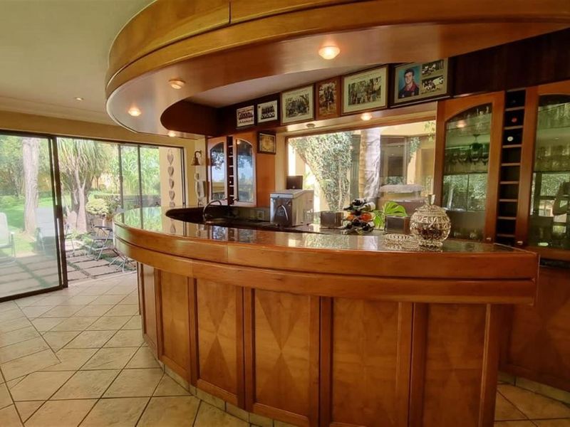 A dedicated bar area featuring a large, curved wooden bar counter with integrated cabinetry, shelving, and recessed lighting, overlooking the garden through sliding glass doors.