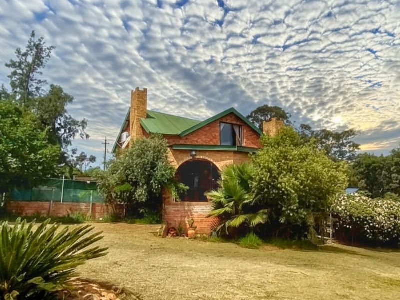 A charming face-brick farmhouse featuring a green metal roof, arched entrance, and established garden surroundings on a spacious rural property.