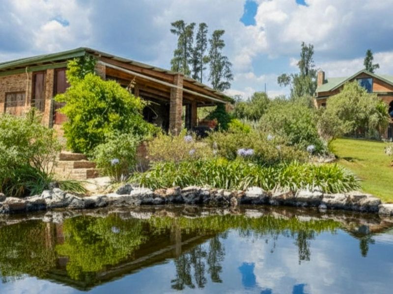 A scenic view of a farm property featuring a natural pond, landscaped garden, and rustic brick buildings with covered patio areas.