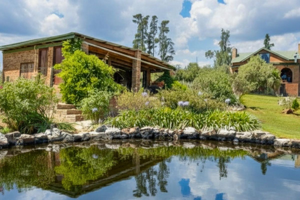 A scenic view of a farm property featuring a natural pond, landscaped garden, and rustic brick buildings with covered patio areas.