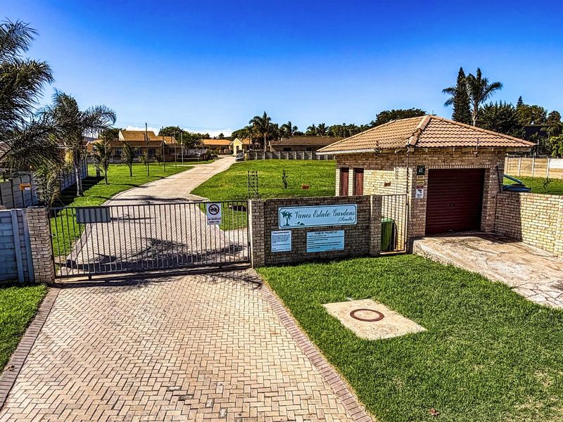 Entrance view of Vanes Estate Gardens featuring a paved driveway, secure metal gate, brick guardhouse, and well-maintained lawn area.