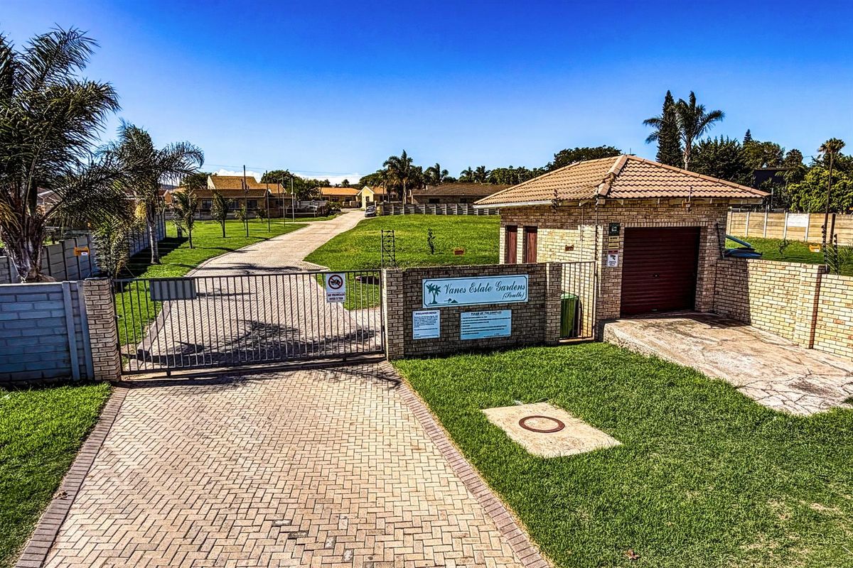 Entrance view of Vanes Estate Gardens featuring a paved driveway, secure metal gate, brick guardhouse, and well-maintained lawn area.