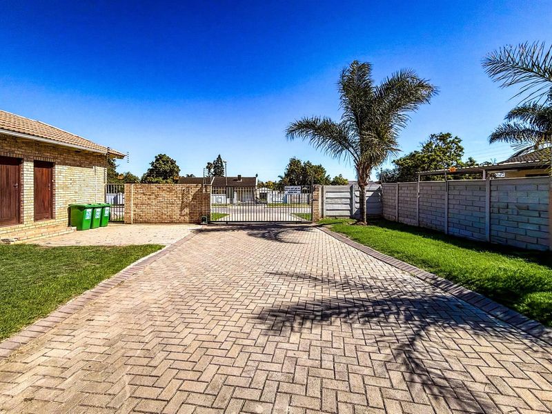 A well-maintained residential property entrance featuring a paved driveway, secure metal gate, brick boundary walls, and established lawn area.