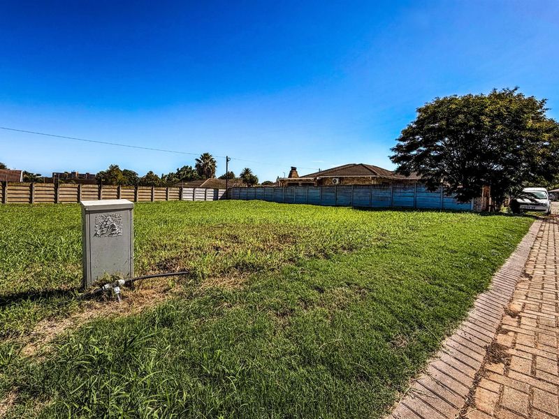 A vacant residential plot featuring a level grass surface, perimeter boundary wall, and an existing electrical connection point.
