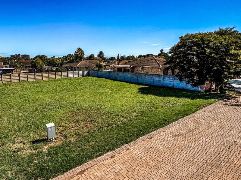 A level, vacant residential plot featuring established lawn, perimeter concrete walling, and paved driveway access.
