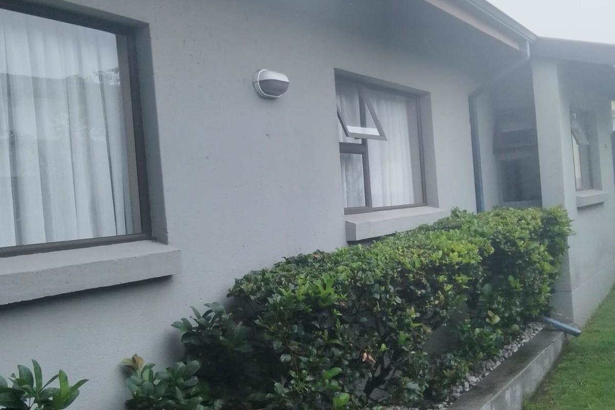 Side exterior view of the house featuring grey rendered walls, aluminium window frames, and a landscaped garden bed with stone mulch.