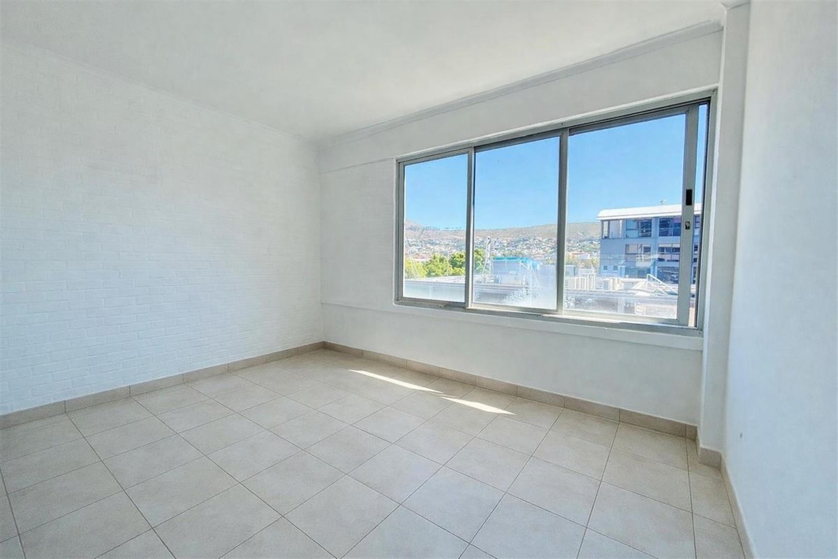 A bright, empty room featuring tiled flooring, white painted brick walls, and large sliding windows offering an urban view.