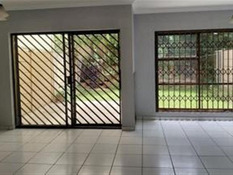 An empty living area featuring tiled flooring, large sliding glass doors with security gates, and window security bars overlooking the garden.