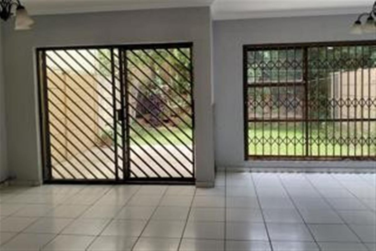 An empty living area featuring tiled flooring, large sliding glass doors with security gates, and window security bars overlooking the garden.