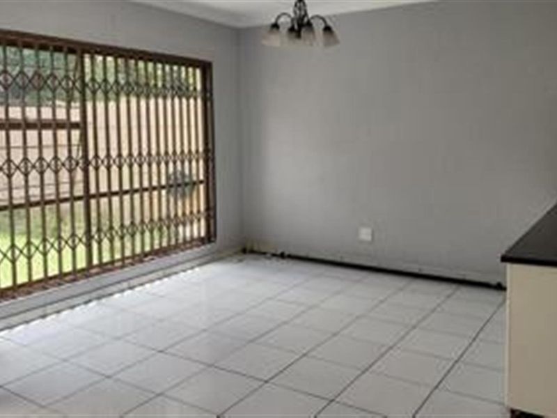 An empty open-plan living area featuring white tiled flooring, a large window with a security gate, and partial kitchen cabinetry.