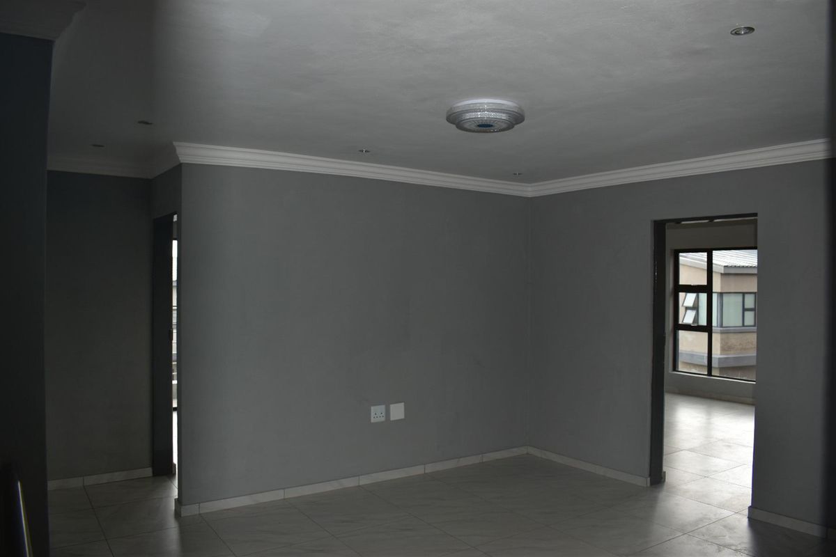 An empty living area featuring neutral grey walls, tiled flooring, and crown molding, with doorways leading to adjacent spaces.