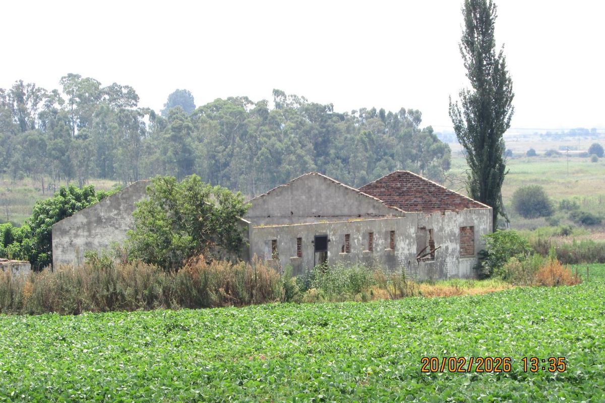A dilapidated farm building structure showing significant deterioration and missing roof elements, situated within a rural agricultural landscape.