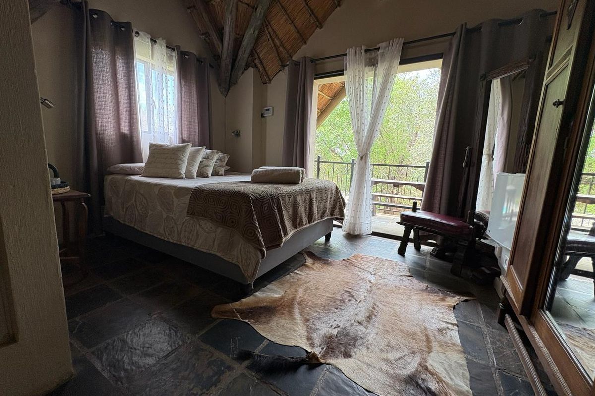 A rustic bedroom featuring a high thatched ceiling with exposed timber rafters, stone tile flooring, and direct access to a private balcony.