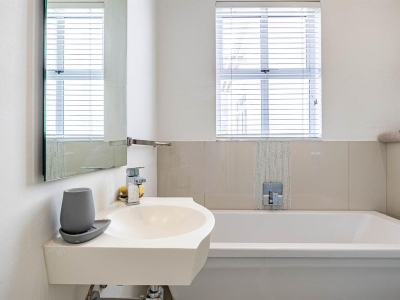 A functional bathroom featuring a wall-mounted basin, built-in bathtub with chrome fixtures, and a tiled splashback with natural light from a window.