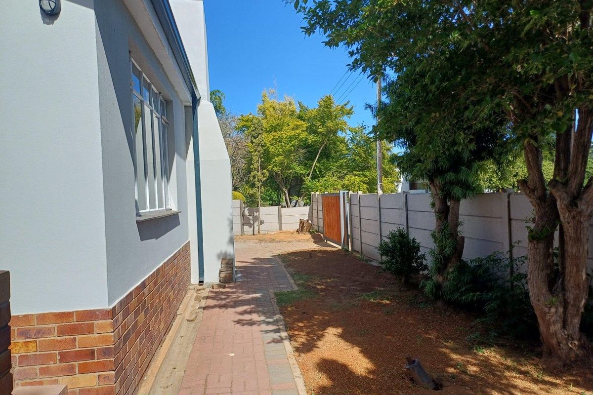 A side exterior view of the property featuring a paved walkway, perimeter walling, and established trees along the boundary.