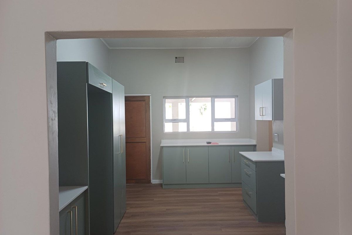 A clean, modern kitchen featuring sage green cabinetry, white countertops, and wood-look flooring, viewed through a doorway.
