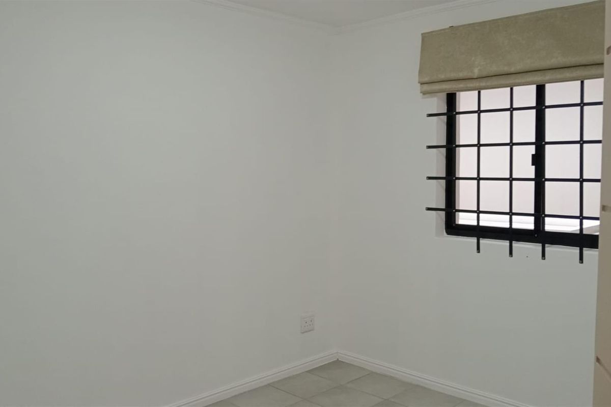 A clean, empty bedroom featuring neutral tiled flooring, white walls, and a window fitted with a security burglar bar and a roller blind.