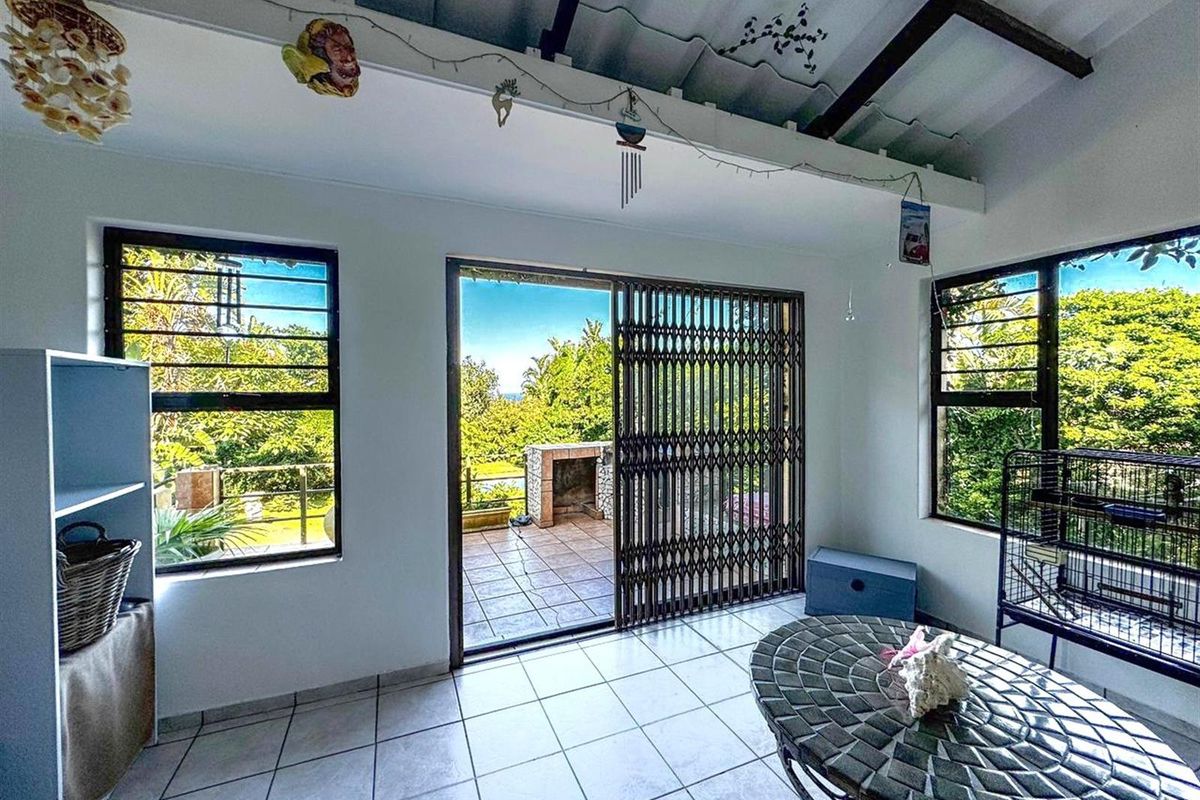 A sunlit living area featuring tiled flooring, large windows, and a sliding security gate leading to an outdoor patio with a built-in braai.