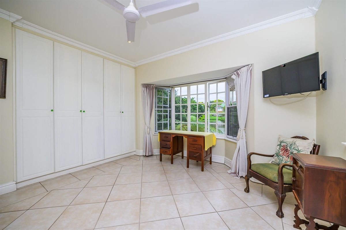 A bedroom featuring tiled flooring, built-in floor-to-ceiling wardrobes, a ceiling fan, and a bay window overlooking the garden.