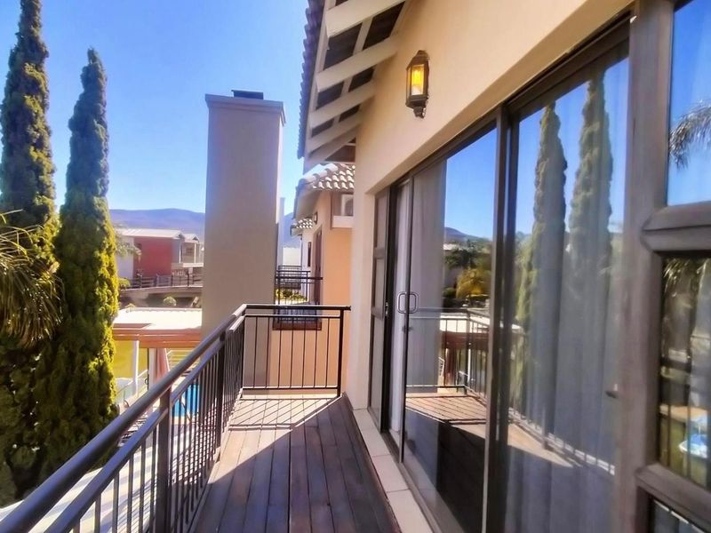 A covered balcony featuring timber decking, metal railings, and large sliding glass doors, offering views of the surrounding estate and water features.