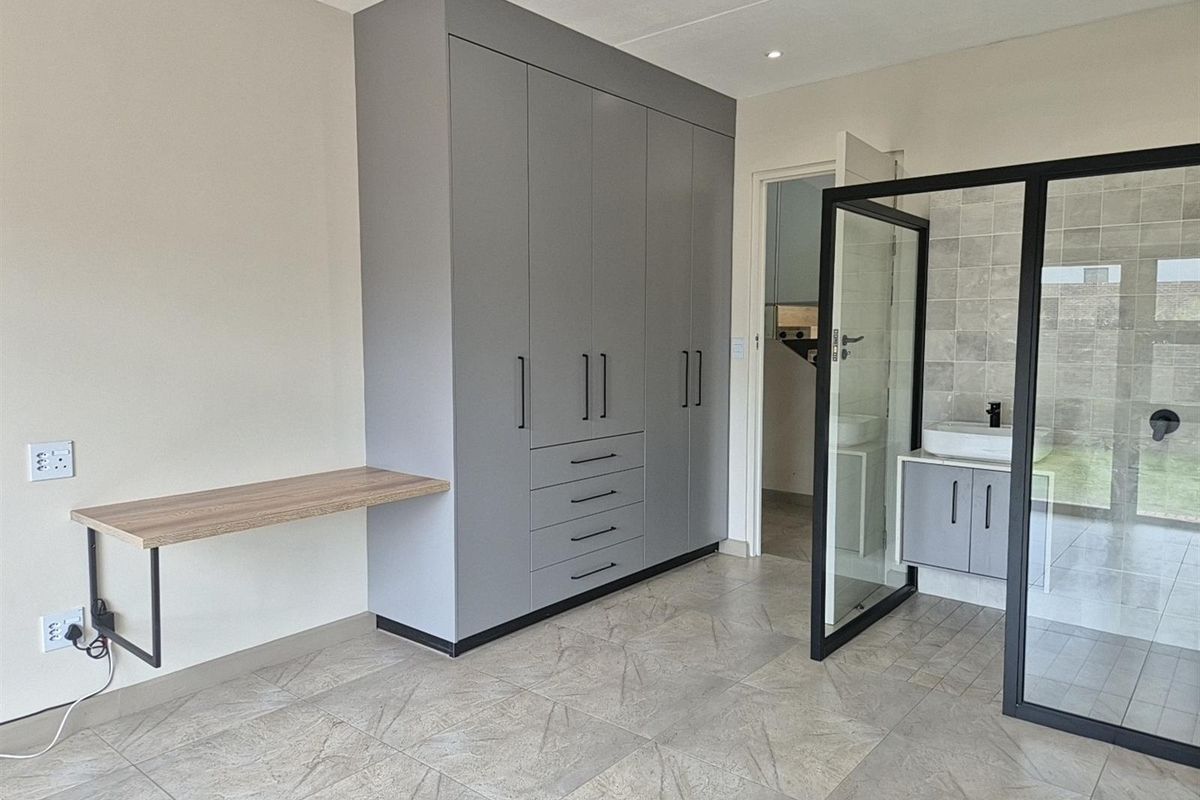 Modern bedroom featuring built-in grey cupboards, a floating desk, and an open-plan en-suite bathroom with a glass shower partition and vanity.