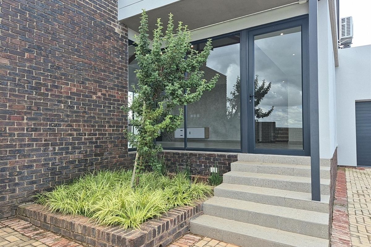 A modern rear exterior view featuring a brick facade, concrete steps leading to a glass door, and a landscaped raised brick planter.