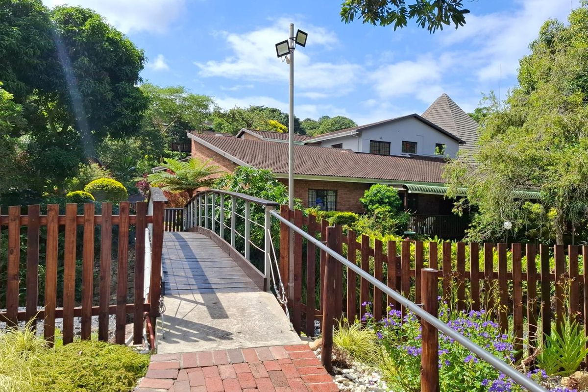A scenic view of a pedestrian bridge leading towards a residential building, surrounded by lush garden landscaping and mature trees.
