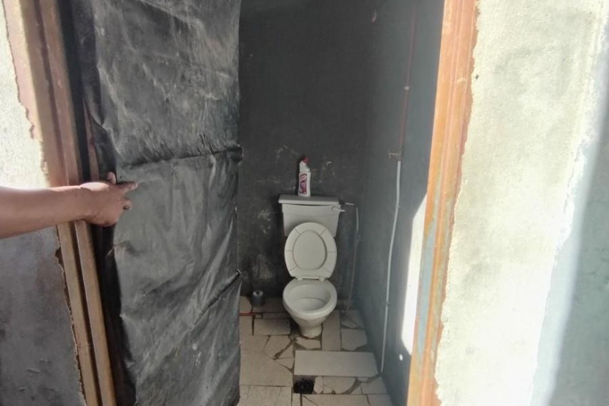 A small, unfinished bathroom featuring a basic toilet, exposed ceiling beams, and damaged floor tiling, requiring significant renovation and finishing work.