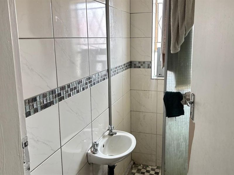 A compact bathroom featuring wall-mounted basin, tiled walls with mosaic accents, and a shower area. The space requires maintenance and cosmetic updates.
