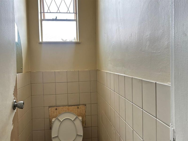 A small, dated bathroom featuring a toilet, partial ceramic wall tiling, and a leaded glass window, requiring cosmetic updates and repairs.