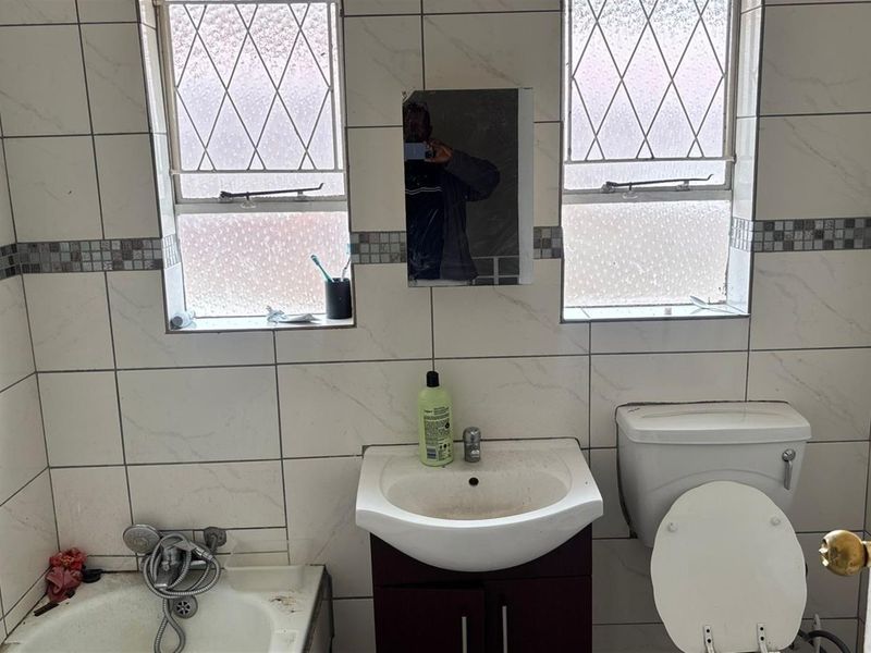 A dated bathroom featuring tiled walls, a bathtub with handheld shower mixer, vanity basin, and toilet, requiring cosmetic updates and deep cleaning.