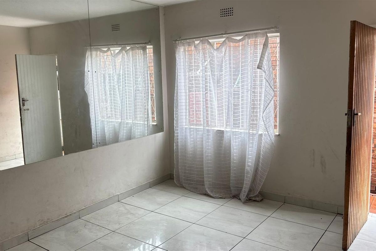 An empty bedroom featuring tiled flooring, a large wall-mounted mirror, and a window with security bars, providing a blank canvas for renovation.