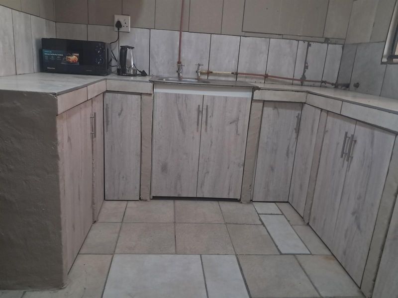 A compact kitchen area featuring light wood-look cabinetry, tiled countertops, and exposed plumbing fixtures requiring finishing touches.
