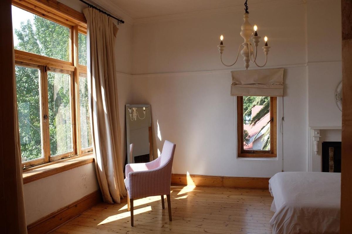 A bright bedroom featuring original timber flooring, large timber-framed windows, a classic fireplace mantel, and a decorative chandelier.