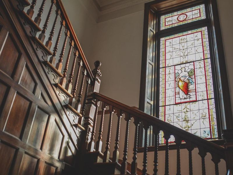 Elegant hallway featuring a grand timber staircase with ornate balustrades and a large, detailed stained glass window providing natural light.