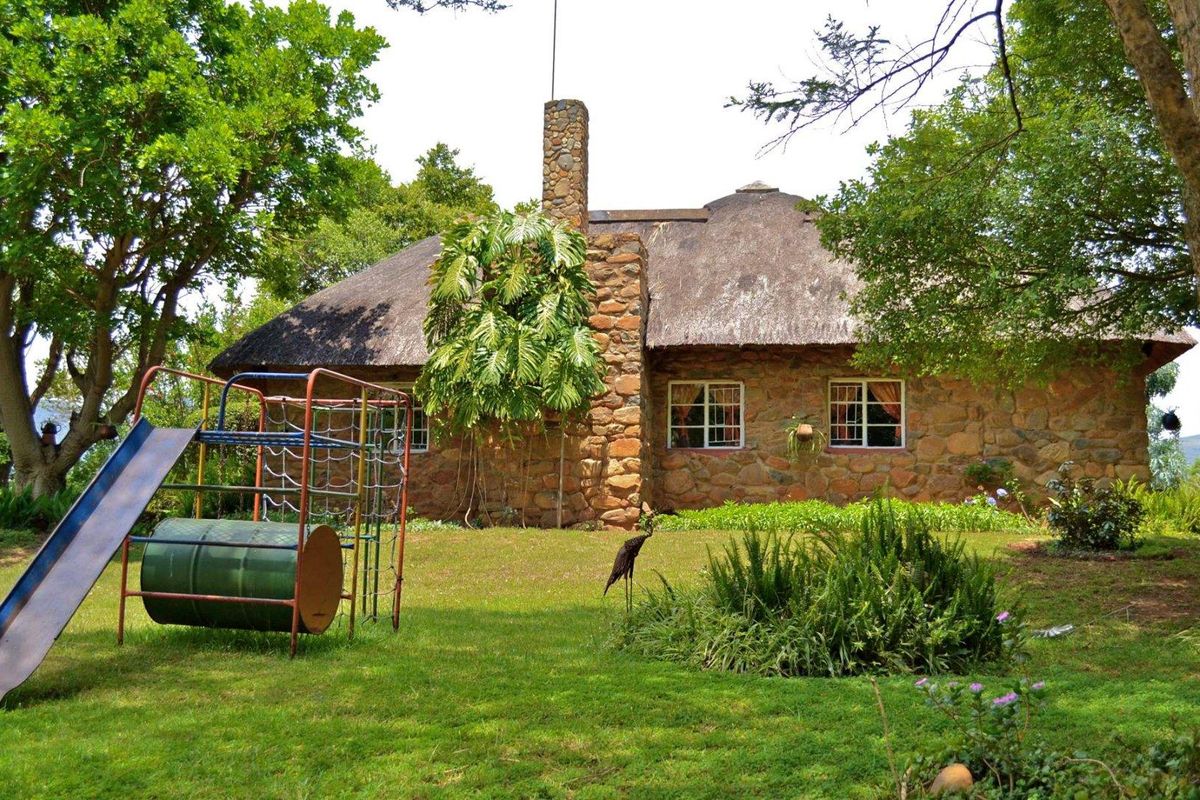 A charming stone-clad farmhouse featuring a traditional thatched roof and a prominent stone chimney, set within a lush, mature garden.