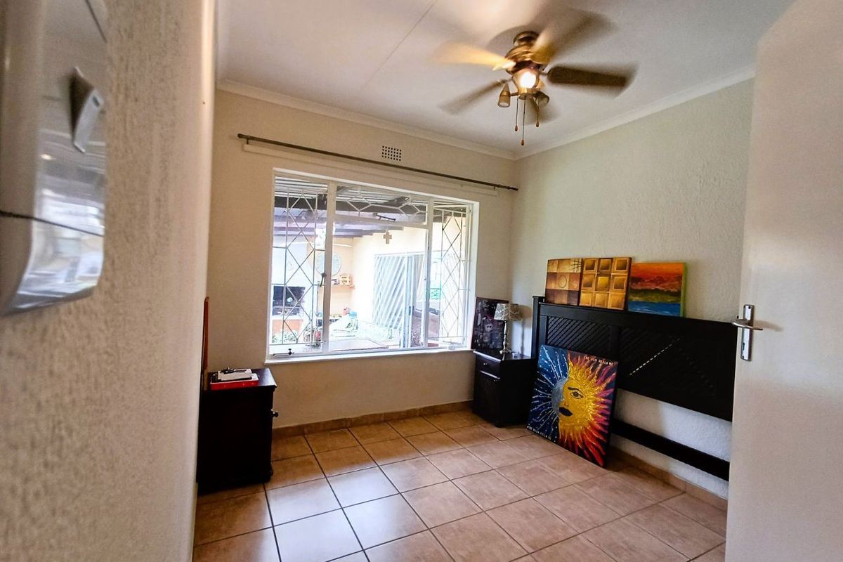 A bedroom featuring tiled flooring, a ceiling fan, and a large window with security bars, offering natural light and a view of the outdoor area.