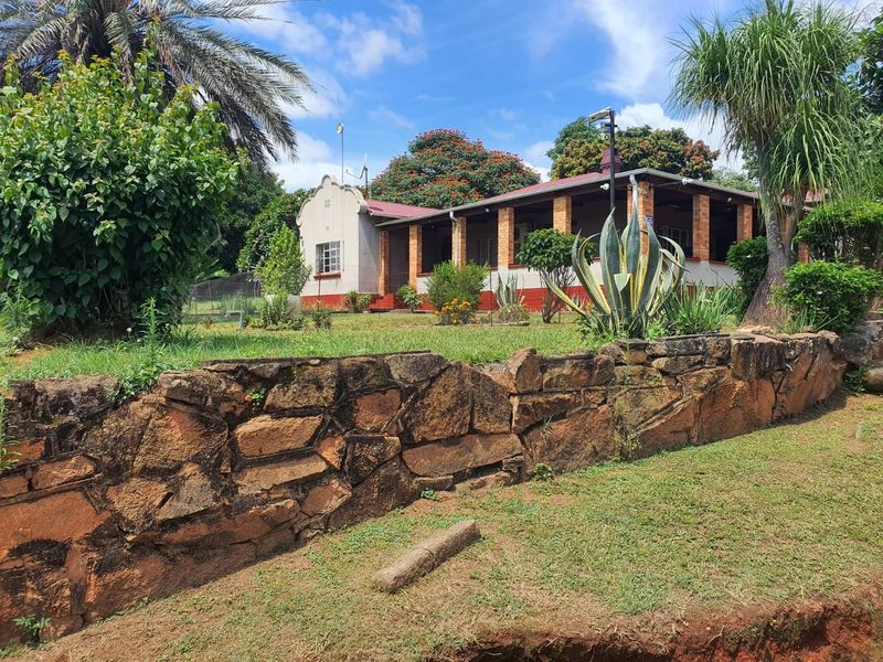 A view of the farm property featuring a traditional house with a covered veranda, set behind a rustic stone retaining wall and landscaped garden.