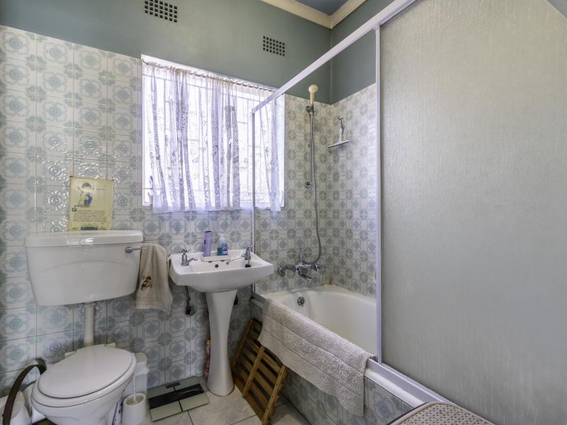 A dated bathroom featuring a pedestal basin, toilet, and a bathtub with a shower mixer and screen, finished with patterned wall tiles.