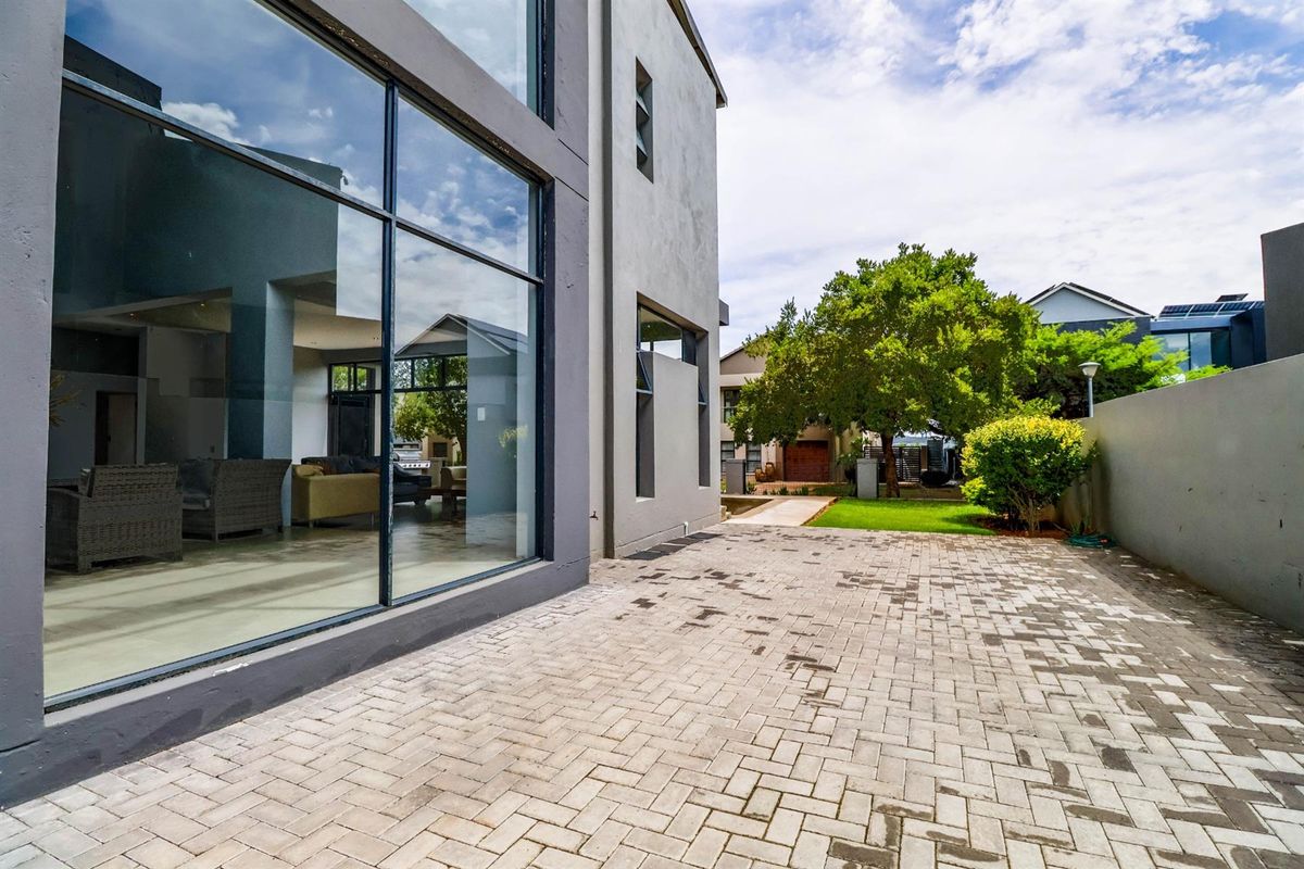 A modern rear exterior view featuring a paved patio area, large floor-to-ceiling glass windows, and a well-maintained garden space.