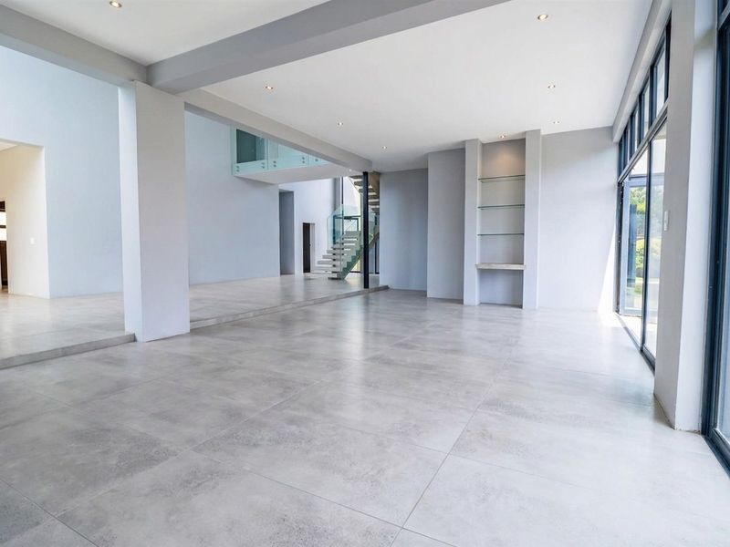 Spacious, modern open-plan living area featuring large format floor tiles, recessed lighting, and a striking glass staircase with floor-to-ceiling glass doors.