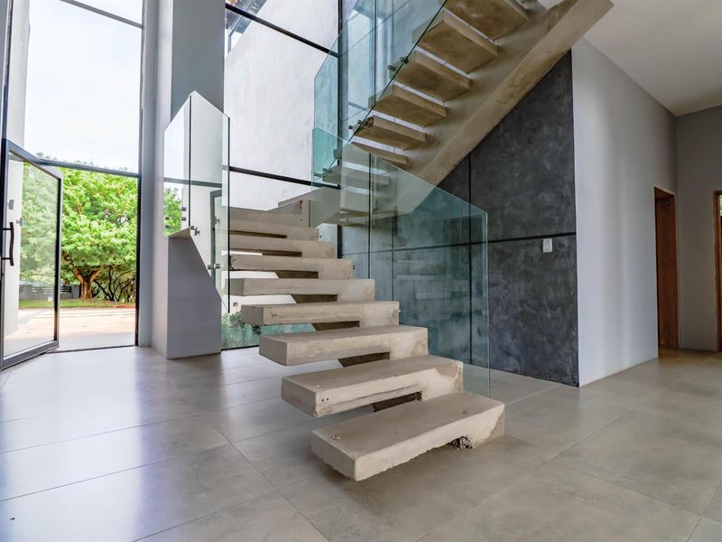 Modern entrance hall featuring a striking floating concrete staircase with glass balustrades, large format floor tiles, and expansive windows providing natural light.