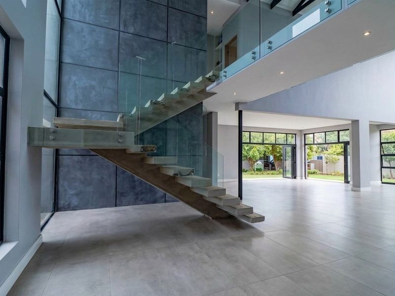 Spacious double-volume living area featuring a modern floating staircase with glass balustrades, large format tiling, and expansive windows opening to the garden.
