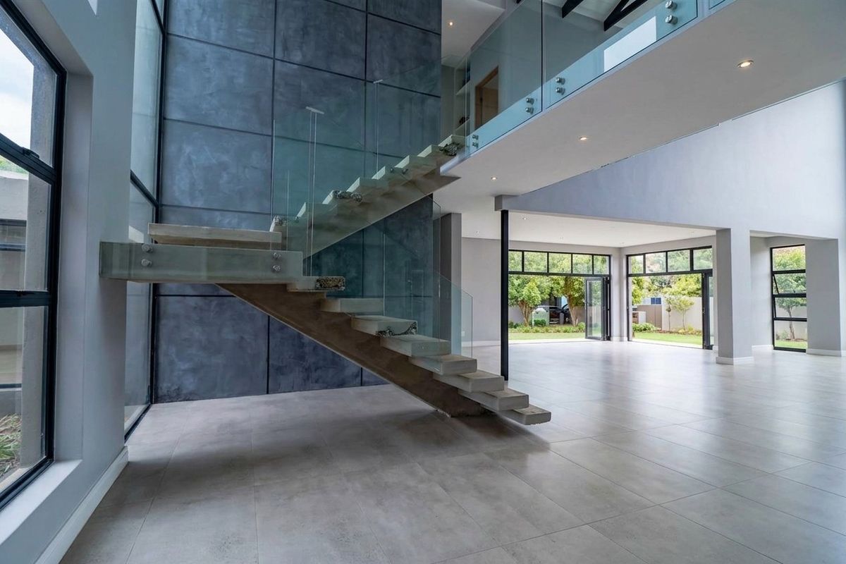 Spacious double-volume living area featuring a modern floating staircase with glass balustrades, large format tiling, and expansive windows opening to the garden.