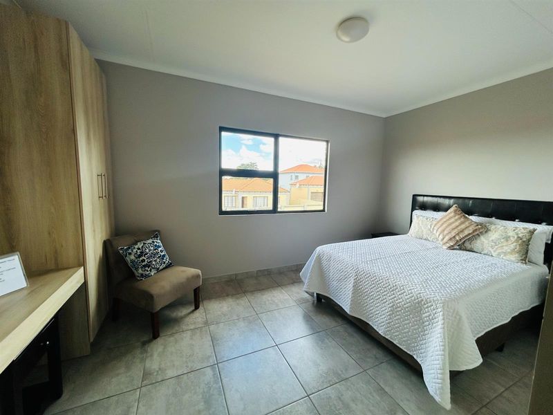 A well-maintained bedroom featuring neutral tiled flooring, built-in wooden cabinetry, and a large window providing natural light and views of the surrounding complex.