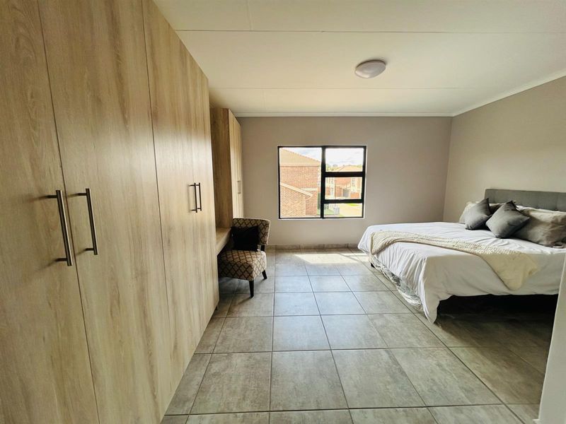 A spacious bedroom featuring light wood-finish built-in cupboards, tiled flooring, and a large window providing natural light and views of the surrounding complex.